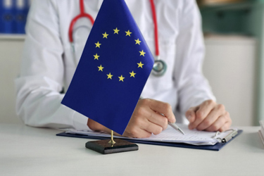 A small European Union flag stands on a desk in the foreground while a doctor in a white coat with a stethoscope writes on a clipboard in a clinical setting, suggesting EU healthcare or medical policy.